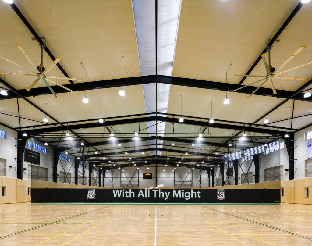 phototonyphillipscom - RMA Sport Indoor gymnasium with a wooden court, overhead lights, black steel beams, and a banner reading 'With All Thy Might'.