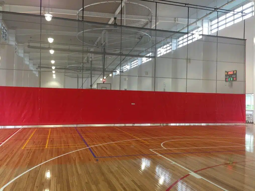 Gallipoli Barracks - RMA Sport Indoor gym with a red divider curtain spanning the court and wooden flooring with white and colored lines for basketball/handball. Behind the curtain, a basketball hoop and netted ceiling fixtures are visible.