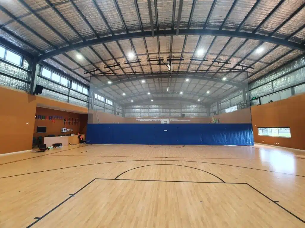IMG_6312 - RMA Sport Indoor gymnasium with a polished wooden floor, blue divider curtain across the back wall, and bright overhead lights in a tall metal ceiling area vignetting windows to the left and back.