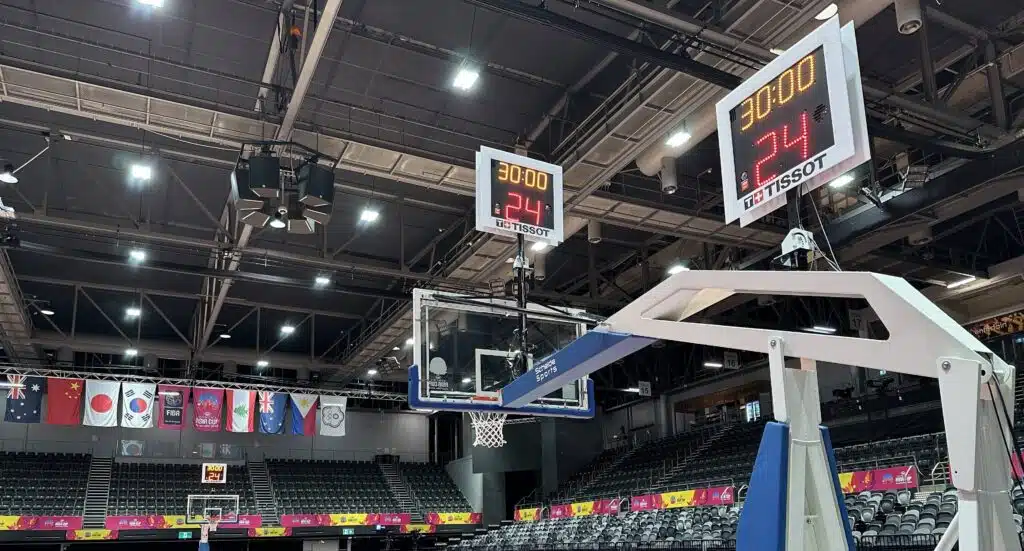 Indoor basketball arena with hanging digital scoreboards showing 30:00 and 24 above empty stands, flags along the lower viewing level.