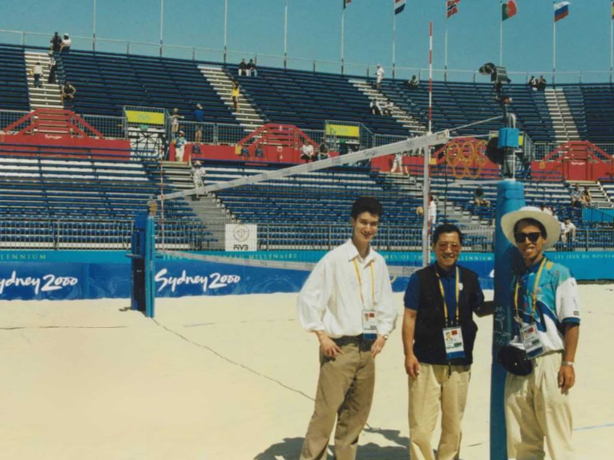 25 Years Website Image - RMA Sport People posing for a photo on a sports arena court with blue bleachers and 'Sydney 2000' banners in the background.