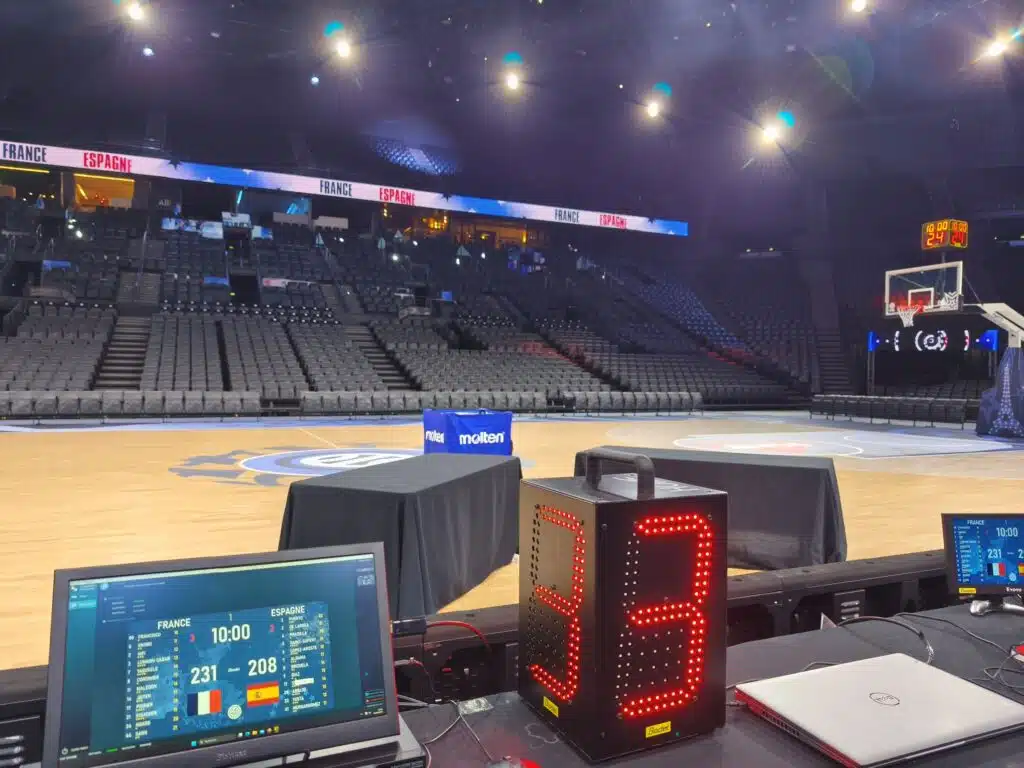 SB-TFD 1 - RMA Sport Basketball arena with bright overhead lights, empty seating, and tech stations with monitors on the sidelines behind a red LED display tower.