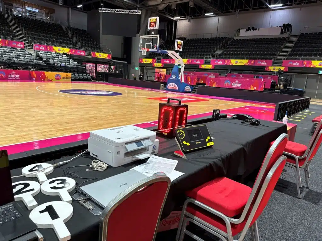 SB-TFD 3 - RMA Sport Basketball arena with empty bleachers, colorful Asia Cup banners along the sidelines, and a scoring table with equipment by the court.