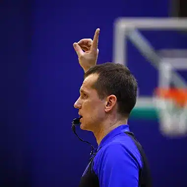 sifflet-electronique-arbitrejpg - RMA Sport Male coach with a whistle in his mouth, raising his index finger during a basketball game on a court.