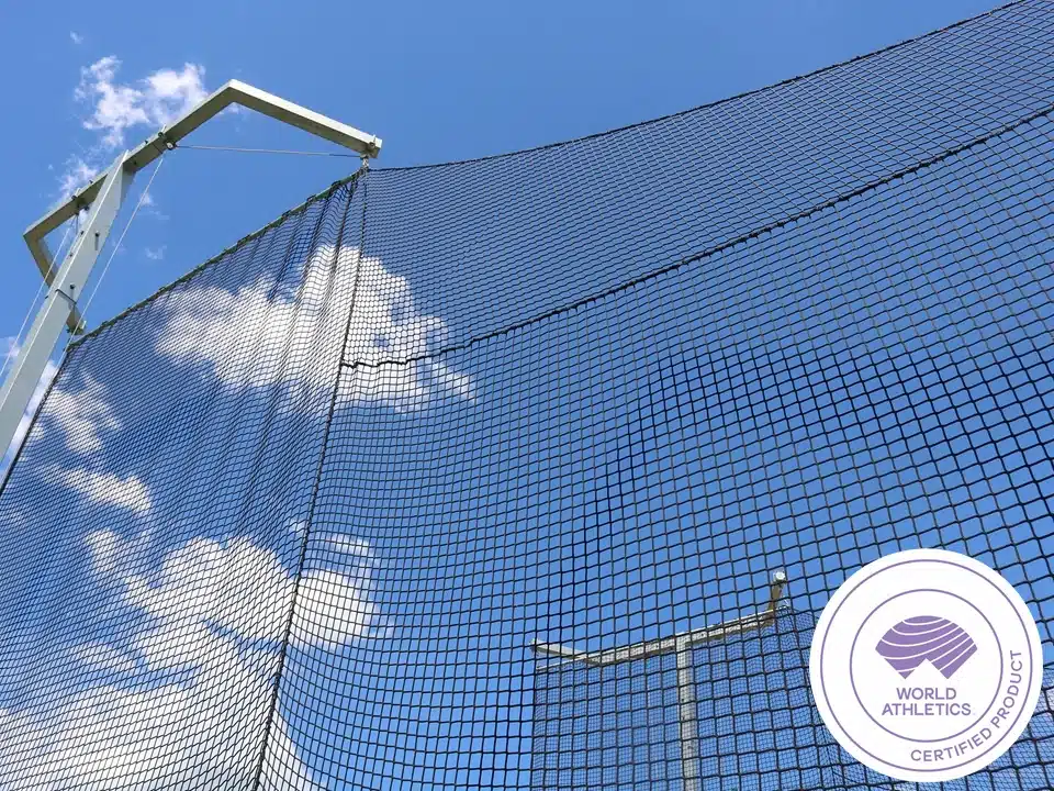 ATH-DTC64 - world-athletics-certified-competition-discus-throw-cage-5 - RMA Sport Chain-link sports fence under a bright blue sky with a metal post and frame; World Athletics certified product badge in the lower right corner.