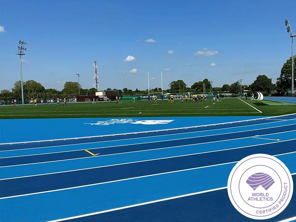 Running Rail Plates - aluminium-track-curbing-3 - RMA Sport Blue running track in the foreground with a green soccer/football field and players practicing in the distance; World Athletics-certified product badge visible in the corner.