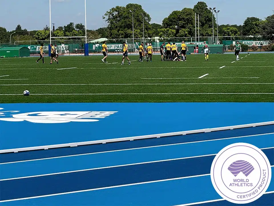 Running Rail Plates - aluminium-track-curbing-4 - RMA Sport Soccer players in yellow jerseys gather in a huddle on a green artificial turf field, with a blue running track in the foreground and a World Athletics badge visible in the corner.