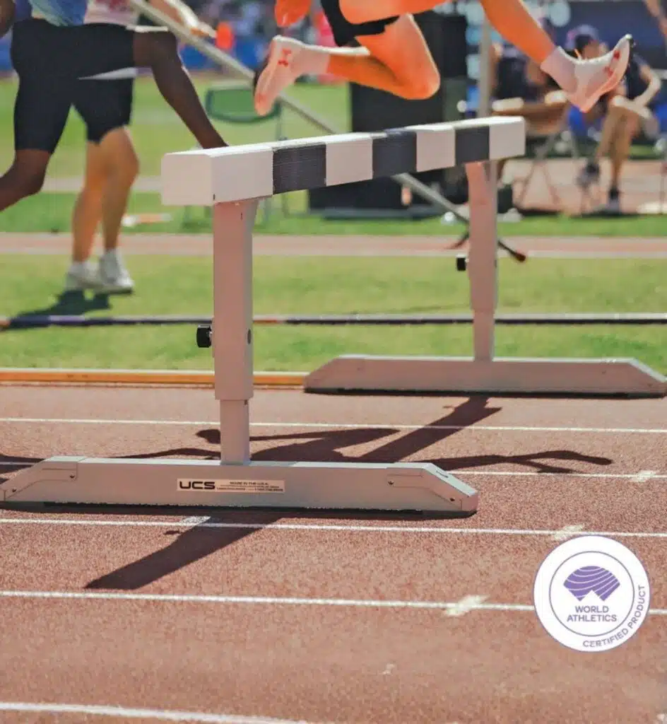 Sprint hurdle on a running track with competitors; foreground shows a white hurdle and a World Athletics Certified product sticker in the foreground.